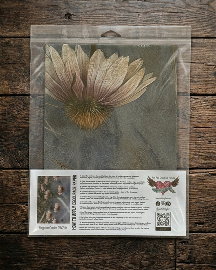 A packaged decoupage paper featuring a large dried flower illustration on a muted background, sealed in a clear plastic bag and placed on a rustic dark wood surface. The lower part of the package includes printed instructions and branding.
