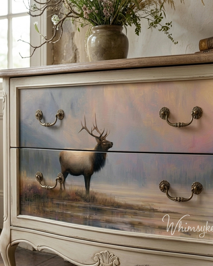 A distressed cream-coloured wooden dresser with two large drawers featuring hand-painted scenic artwork of a Stag standing at the waters edge under a soft pastel sky. The dresser has ornate metal drawer pulls and is styled in a room setting with a window, vase, and books.