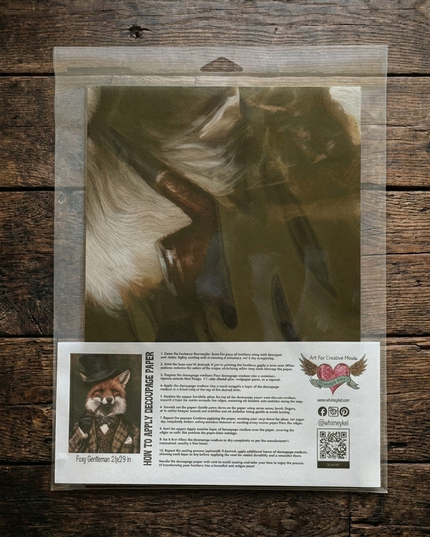 A packaged decoupage art print of a fox wearing a bow tie, displayed on a rustic dark wood background. The print is sealed in a clear plastic bag with a printed instruction card.